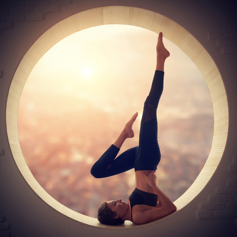 Beautiful sporty fit yogi woman practices yoga handstand asana Salamba Sarvangasana - shoulderstand pose in a round window with arial view of the city at sunset.